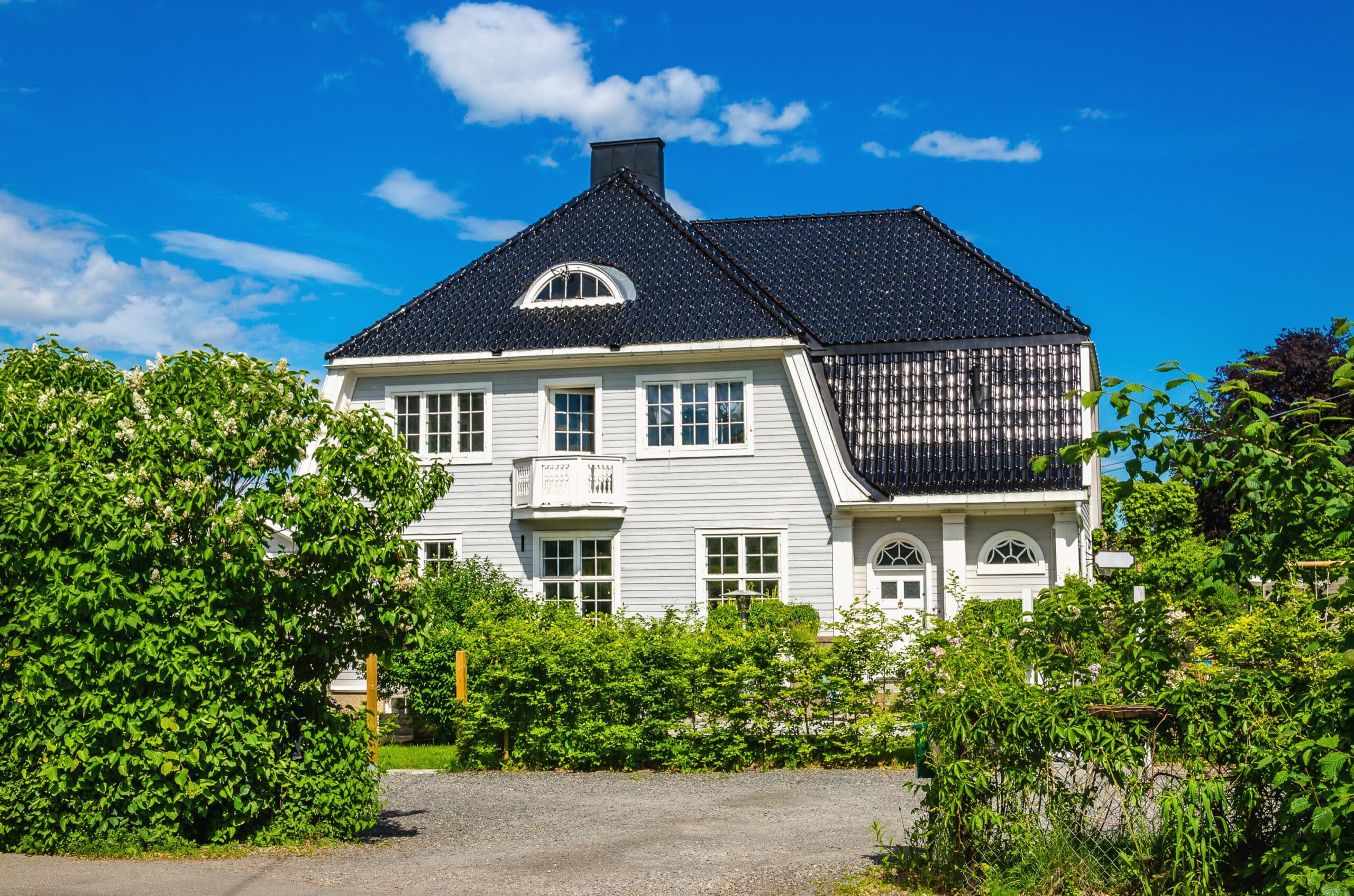 Stor hvit enebolig med mørkt tak og symmetrisk fasade omgitt av grønn hage og busker på en solrik dag. Bildet viser boligen sett rett forfra og gir et tydelig inntrykk av eiendommens størrelse, stil og velstelte uteområde.